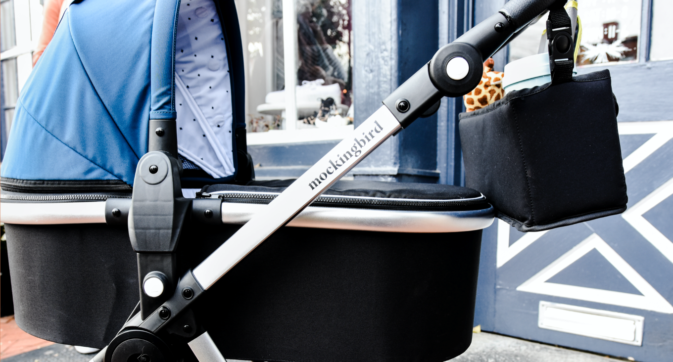 Close-up of a blue Mockingbird stroller with a black cup holder attached, parked outside near a building with a window and door.