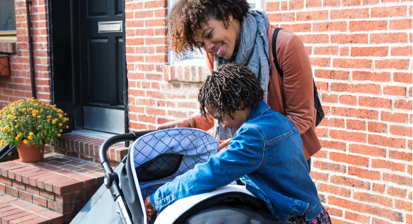 An adult and a child look into a stroller together outside a brick building, with potted flowers visible on nearby steps.