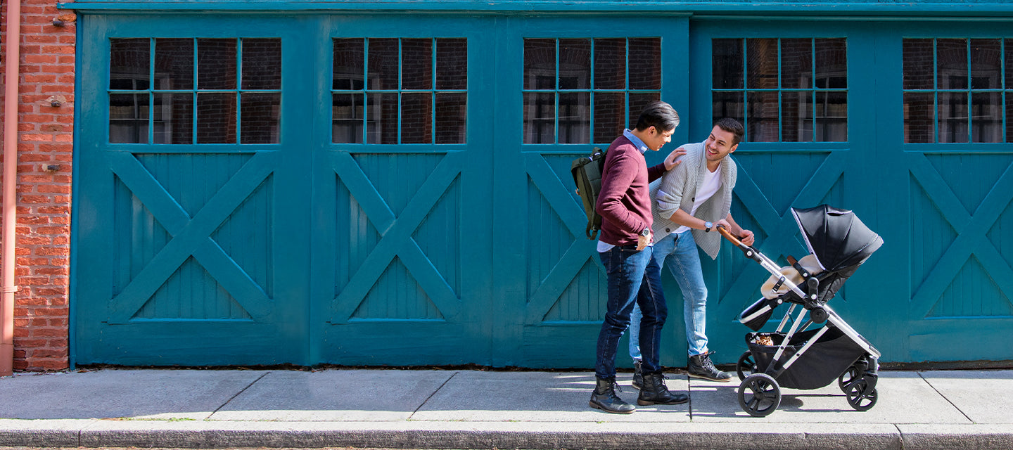 Two men chatting beside a stroller on a city sidewalk, with a large green door in the background.