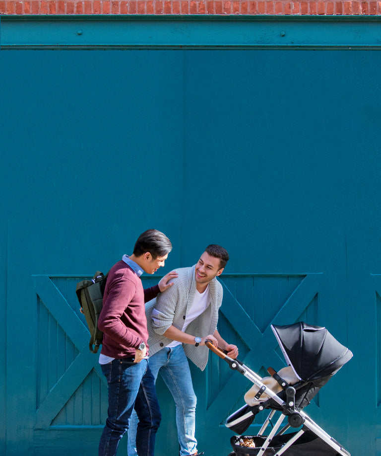 Two men with a baby stroller chatting happily in front of a teal backdrop.