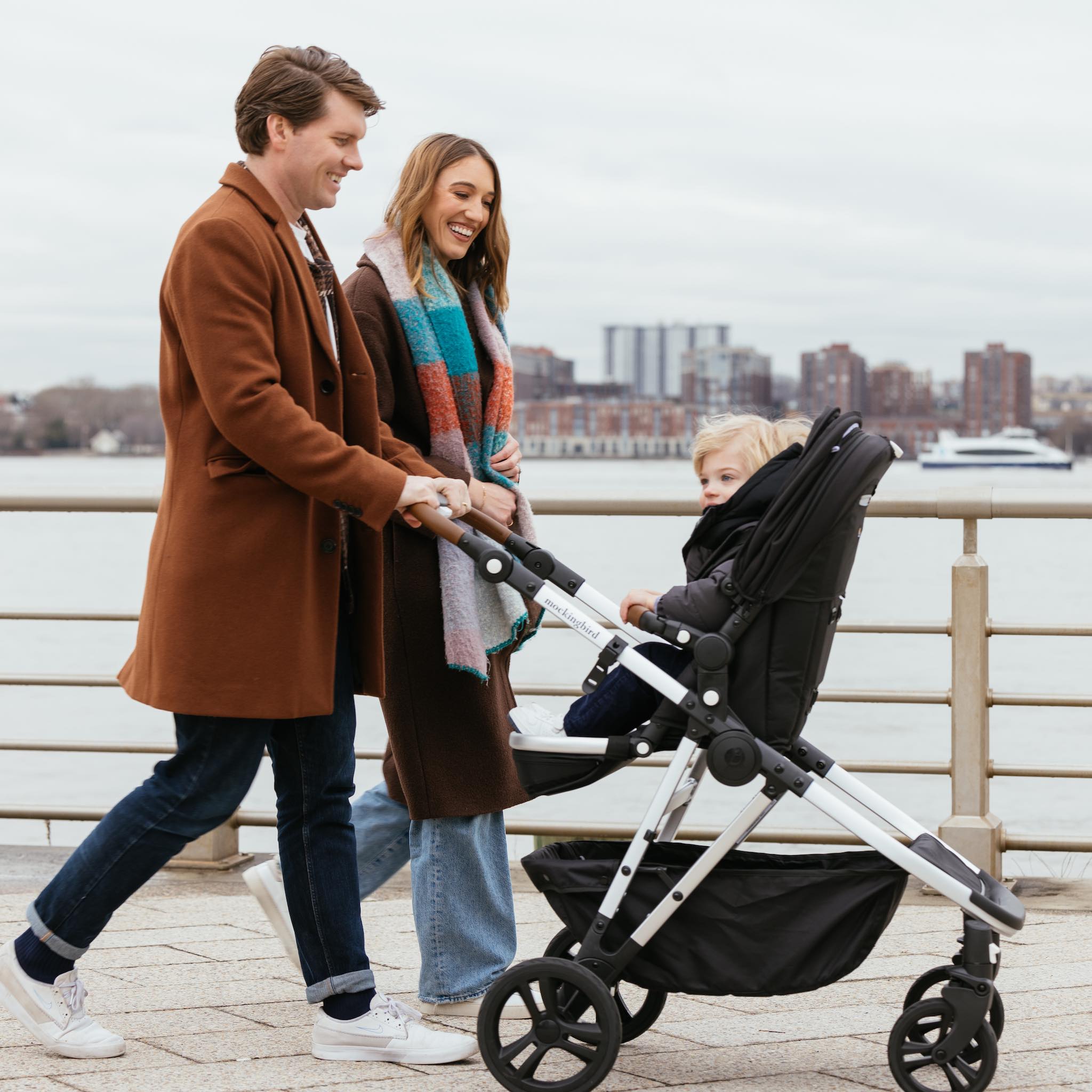 A couple walking with their young child in a stroller by a waterfront promenade, smiling and holding hands.