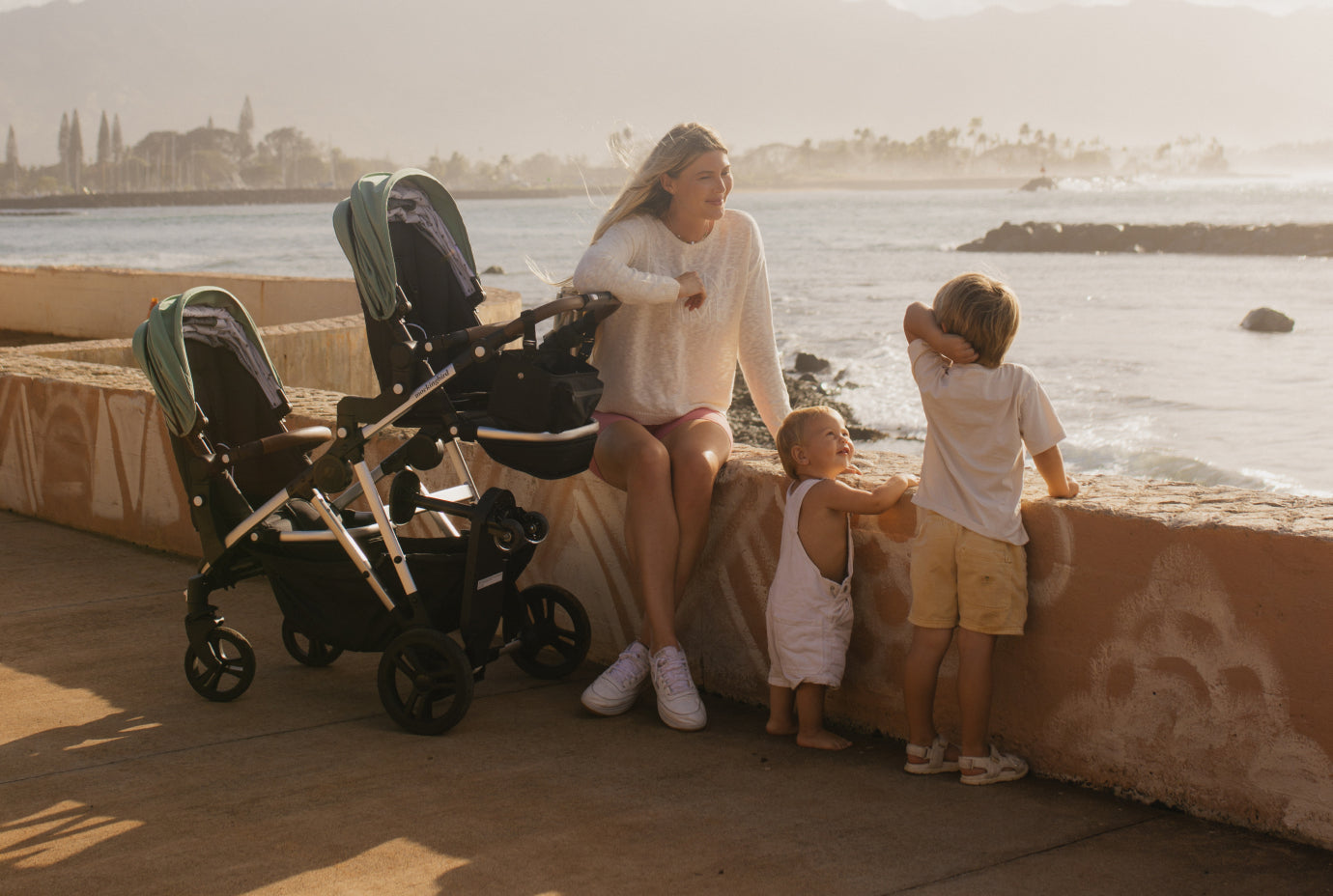 A woman with a stroller sits on a beachside promenade, accompanied by two young children as they look out towards the ocean.
