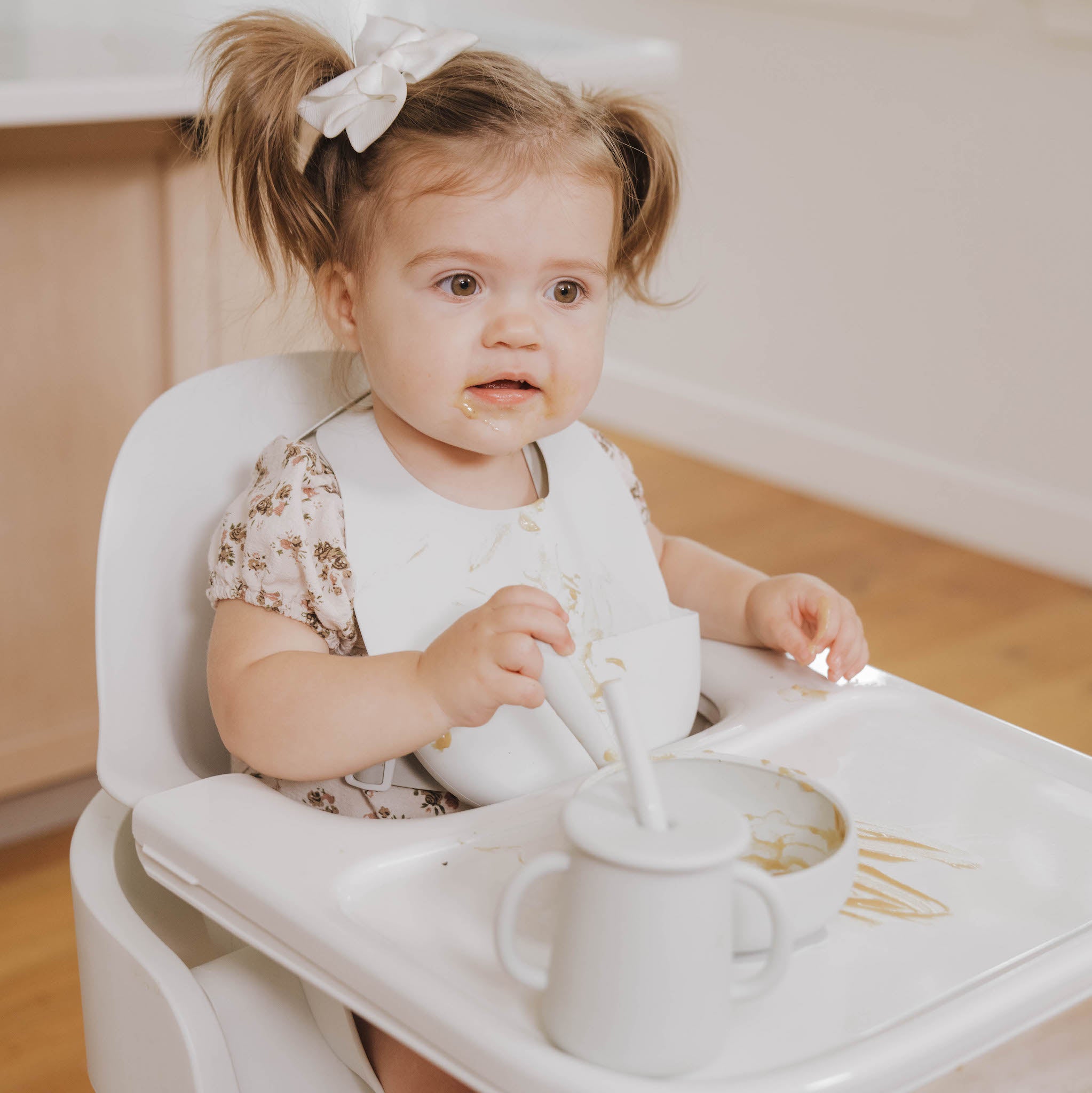 Toddler girl with pigtails eating messily in a high chair, holding a spoon with food on her face and bib.