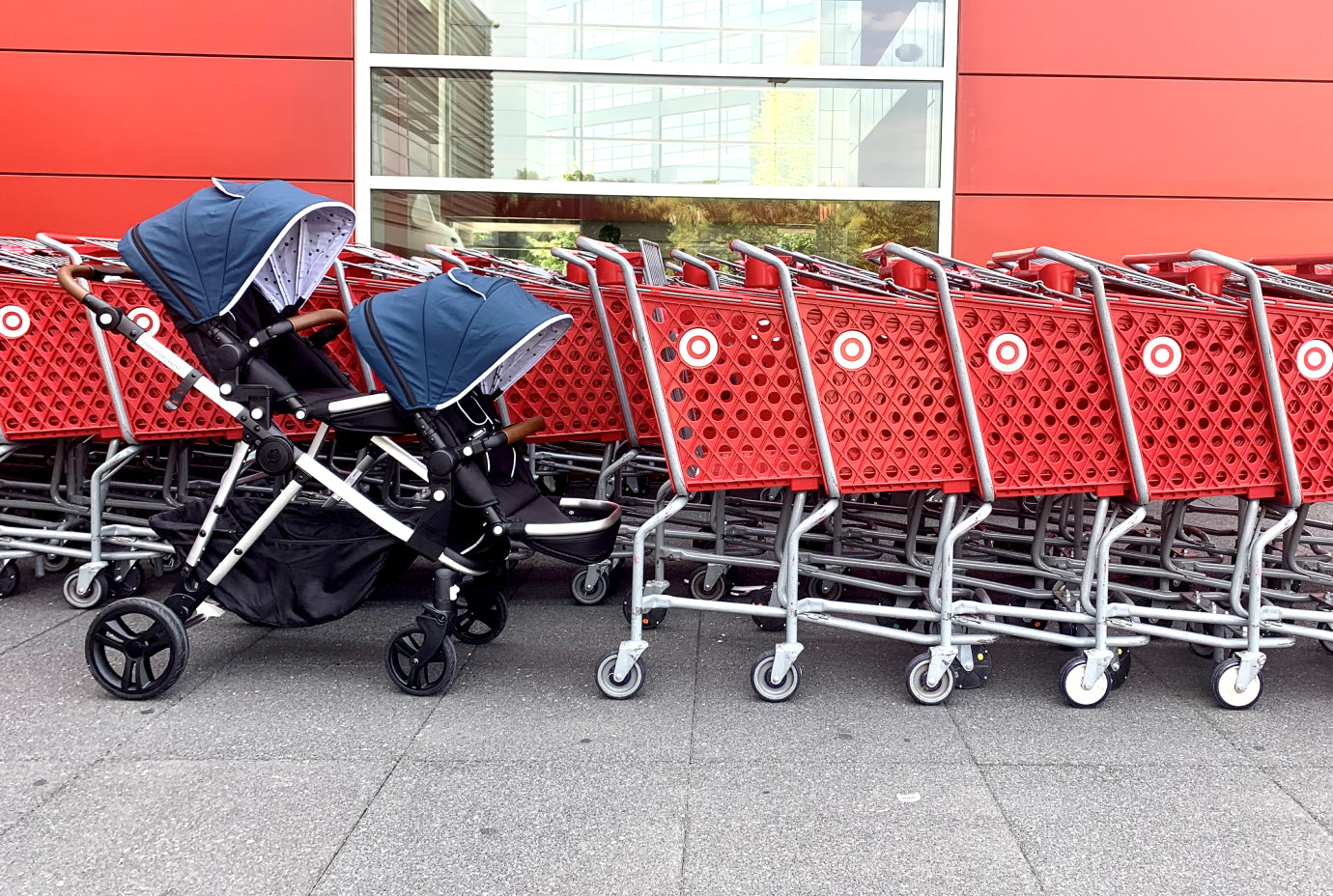 Double stroller parked outside next to a row of red shopping carts against a red and glass building facade.