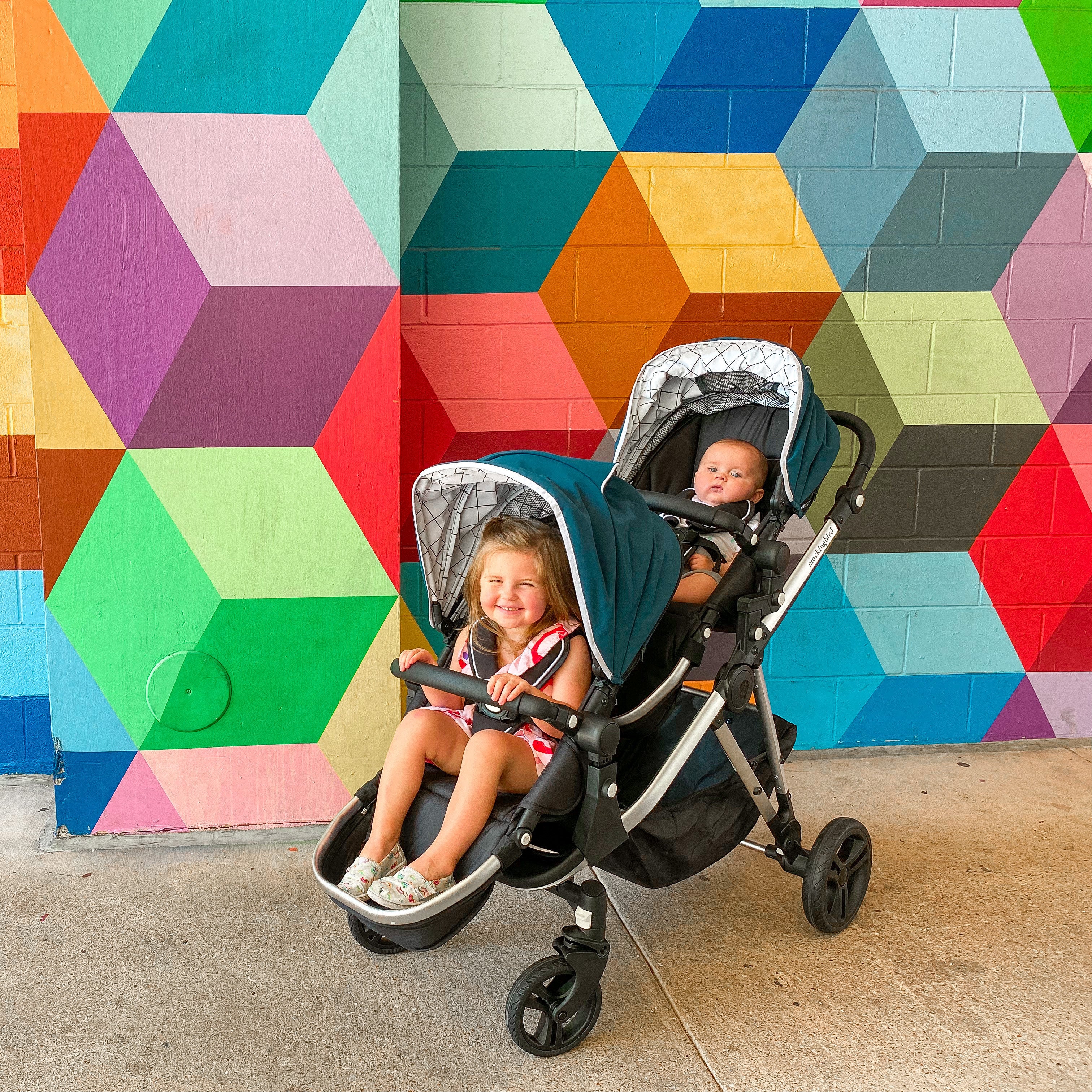 Two children in a double stroller in front of a colorful geometric mural. one child smiles while the other appears to be asleep.