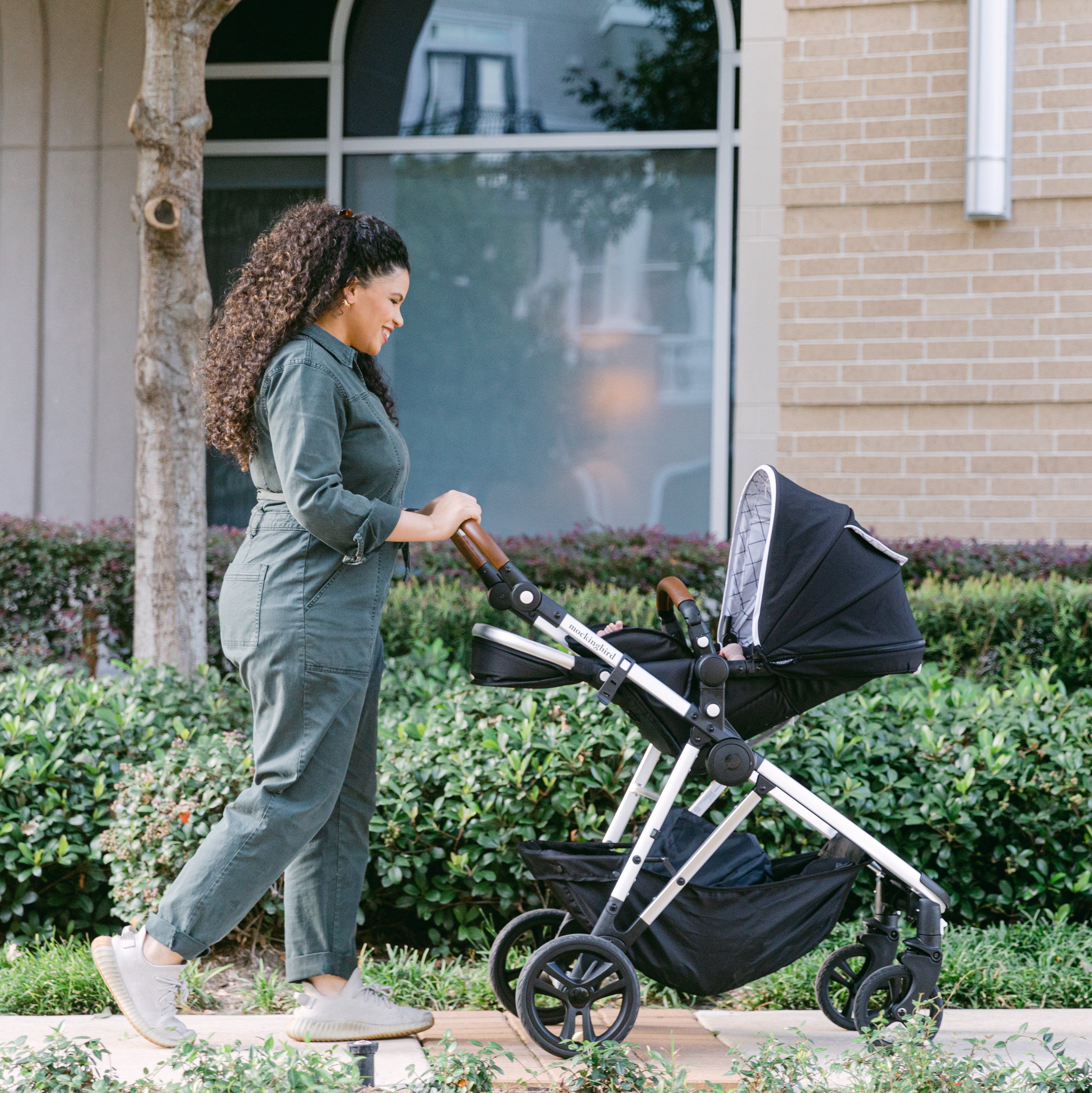 A woman in a green jumpsuit pushing a baby stroller along a sidewalk next to a building with large windows and green shrubbery.