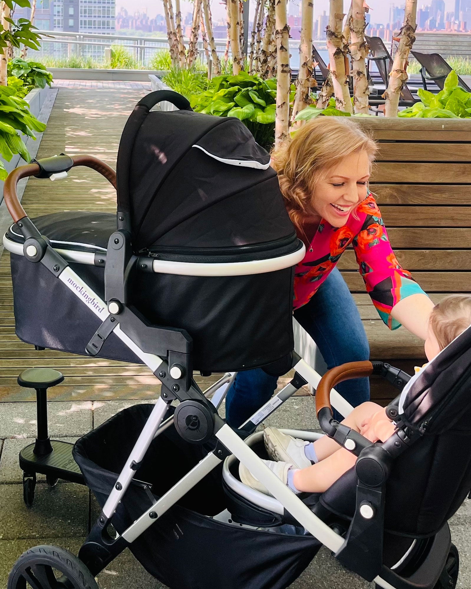 A woman in a colorful blouse smiles at a child in a black mockingbird-prod stroller outside.