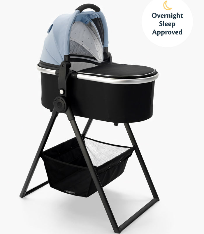 A mockingbird-prod bassinet with a blue canopy on a black bassinet stand, featuring a storage basket underneath and a label indicating "overnight sleep approved. #color_sky