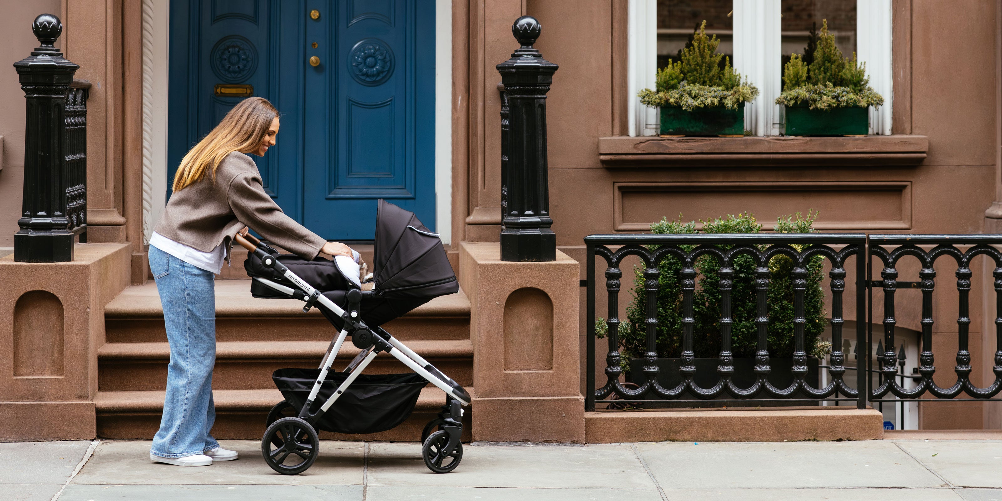 Woman pushing a stroller past brownstone houses with colorful doors on a city street.
