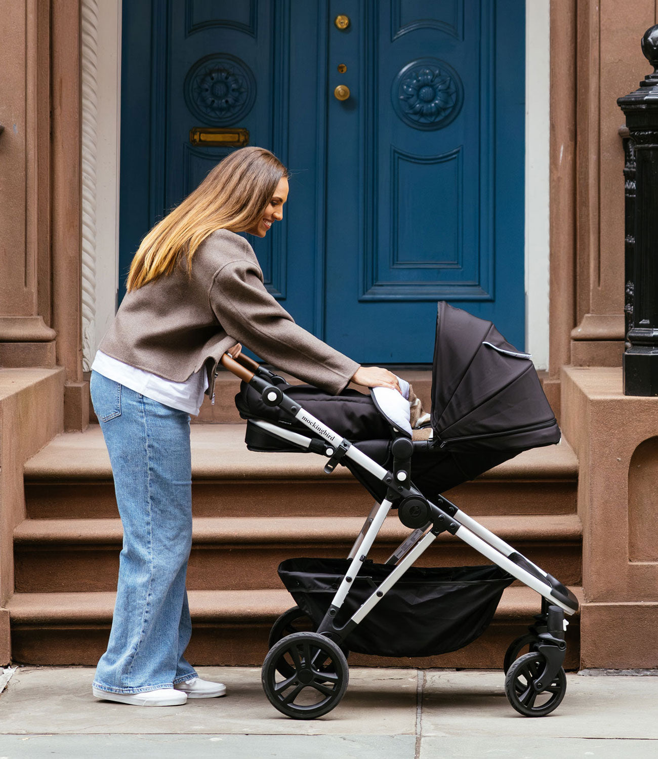 A woman in a brown jacket and jeans pushing a baby stroller near a blue door on a city street.