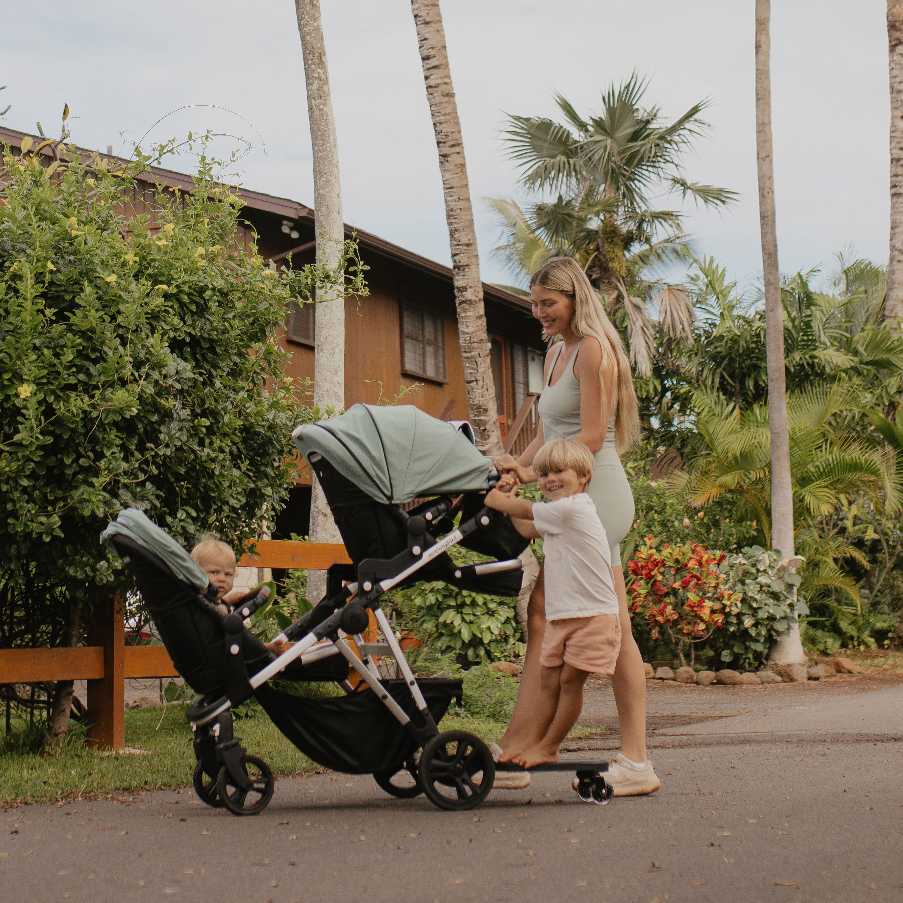 A woman strolls outdoors with a baby in a stroller, accompanied by a young boy on a skateboard, in a tropical setting with palm trees.