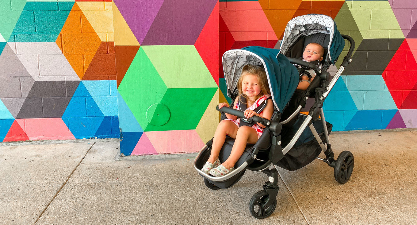 Two children in a stroller in front of a colorful wall.