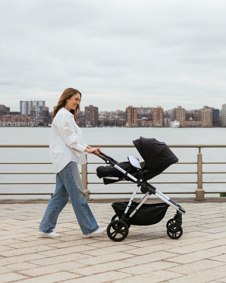 A woman walking along a riverfront promenade, pushing a black stroller. she is dressed in a white shirt and blue jeans, with an urban skyline in the background.