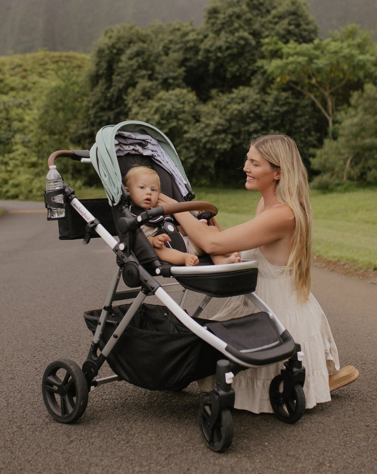 A woman in a white dress smiles while pushing a baby in a stroller along a scenic pathway surrounded by greenery.