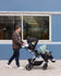 A young man walks outdoors pushing a double stroller with two young children, past a blue building.