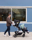 A young man walks outdoors pushing a double stroller with two young children, past a blue building.