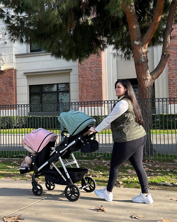 A woman walks outdoors pushing a double stroller with two infants, near a tree and a fence, in casual attire.