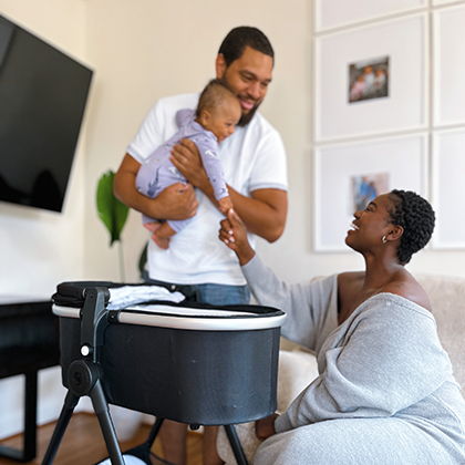 A father holding a baby stands facing a mother seated beside a bassinet in a cozy living room.
