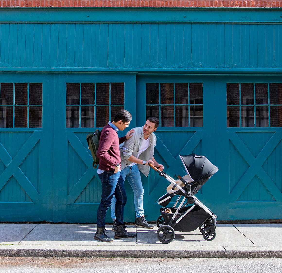 Two men, one pushing a stroller, talking and smiling on a city sidewalk in front of a teal building with large doors.