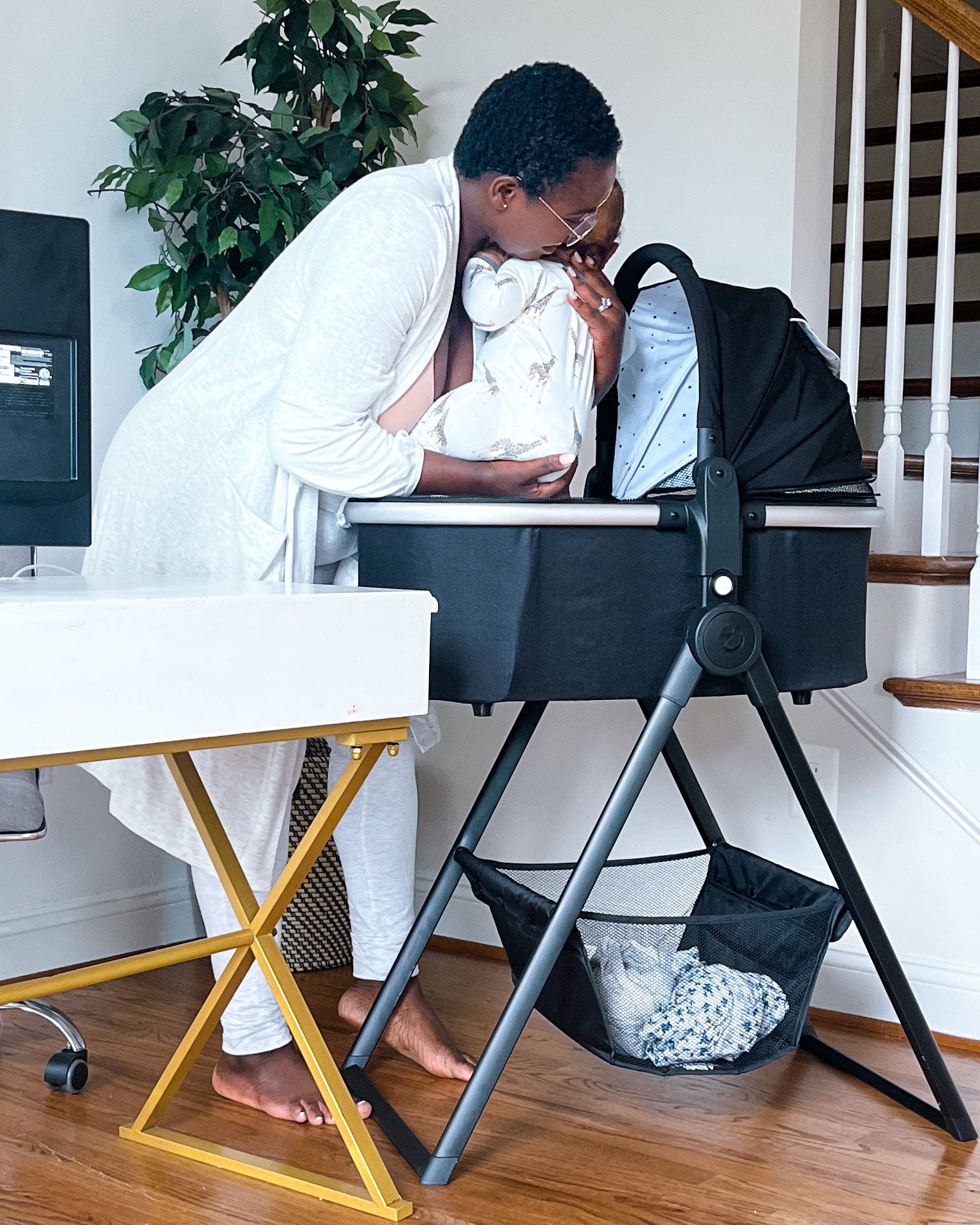 A woman in a white robe kisses a baby cradled in a mockingbird-prod bassinet stand while standing at a home office desk with a computer and a plant.