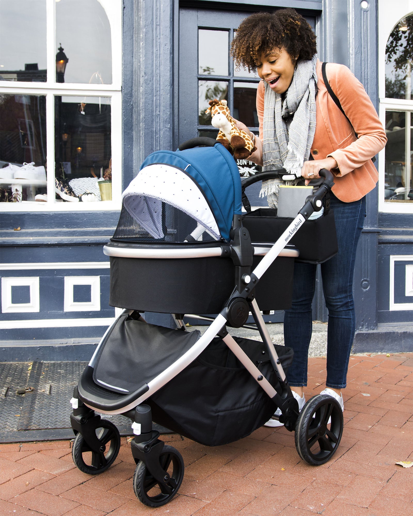 A woman smiling at a baby in a mockingbird-prod Stroller outside a shop, holding a toy giraffe.