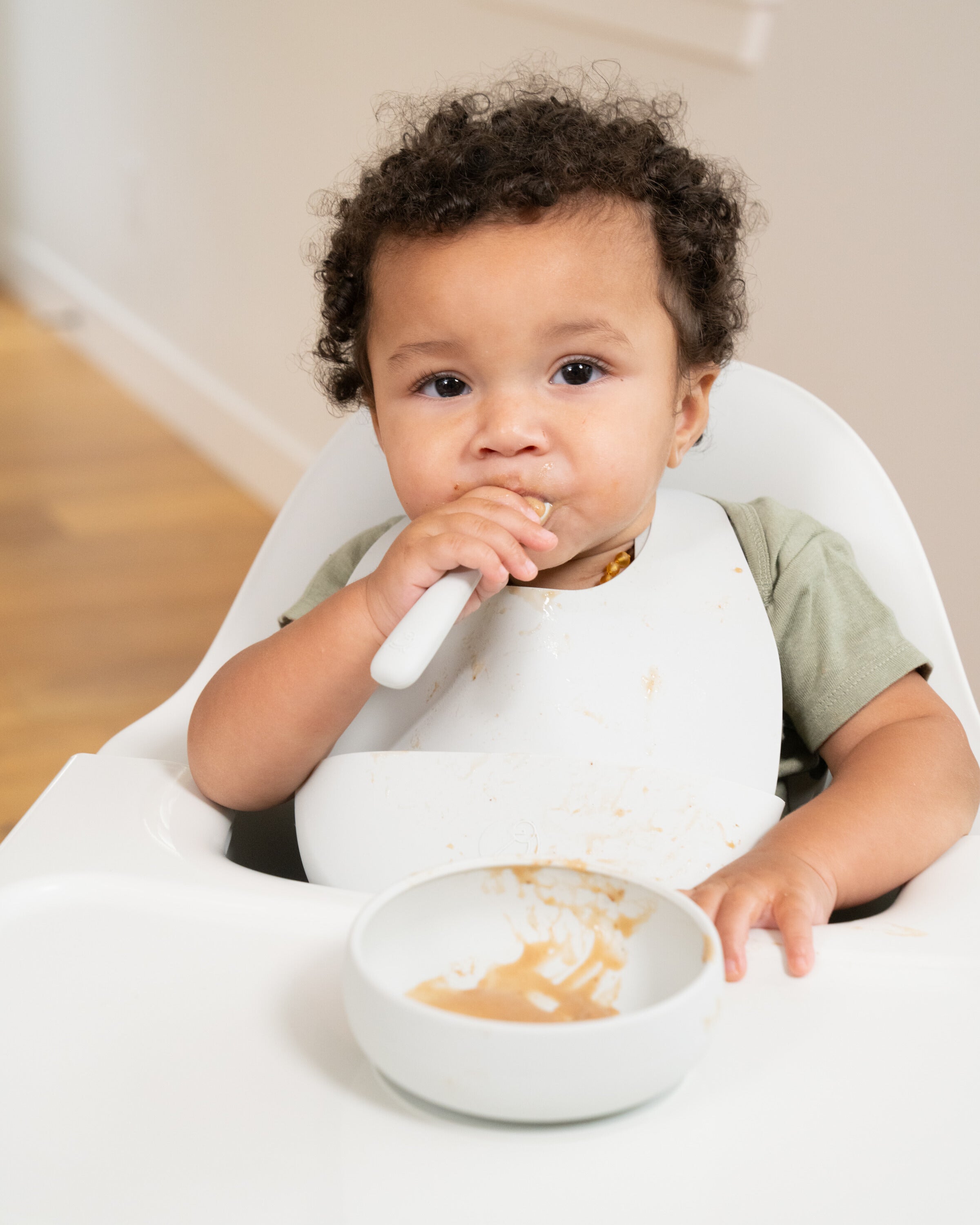 A small child  with brown curly hair enjoying a meal while sitting in the Mockingbird High Chair