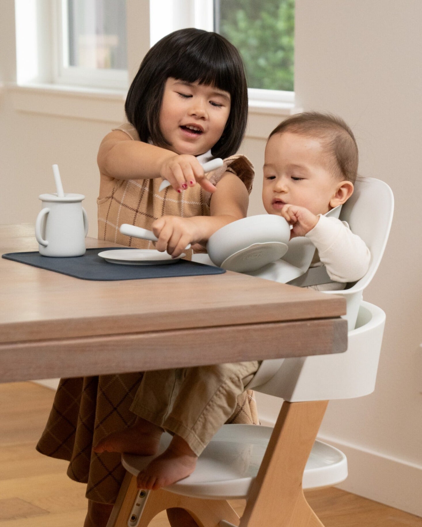 Older child and smaller child at the kitchen table enjoying a meal. The older child is using the Mockingbird dishware to feed her little brother who is sitting in the Mockingbird High Chair