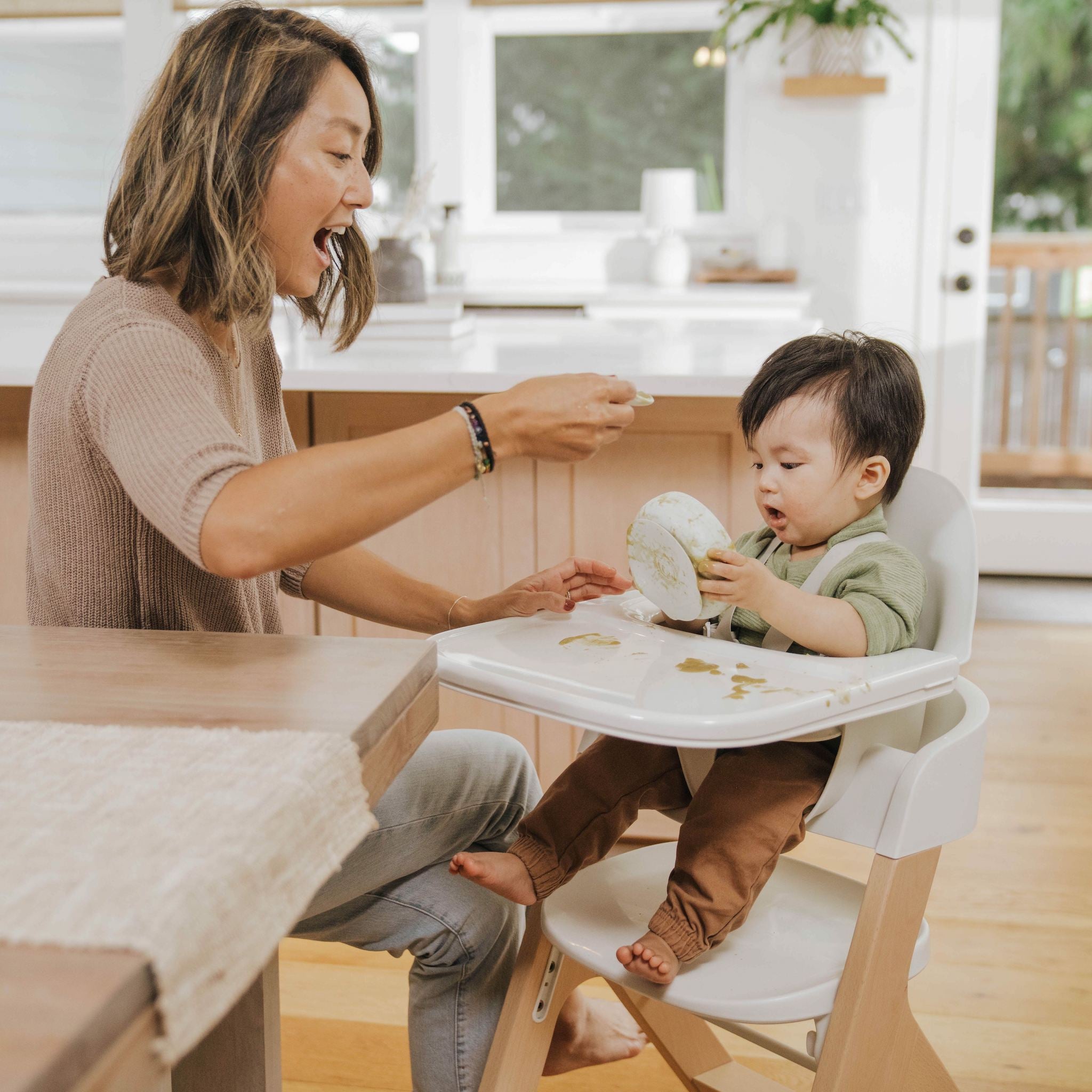 A mother feeding her toddler in a high chair, both smiling as the child attempts to eat by himself.